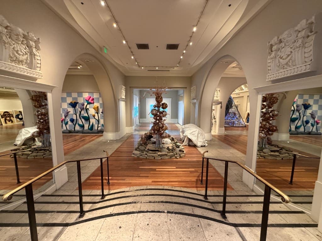 Inside the Coliseum of Art, with arched galleries, glass flowers, warm wood floors, and sculptural light.