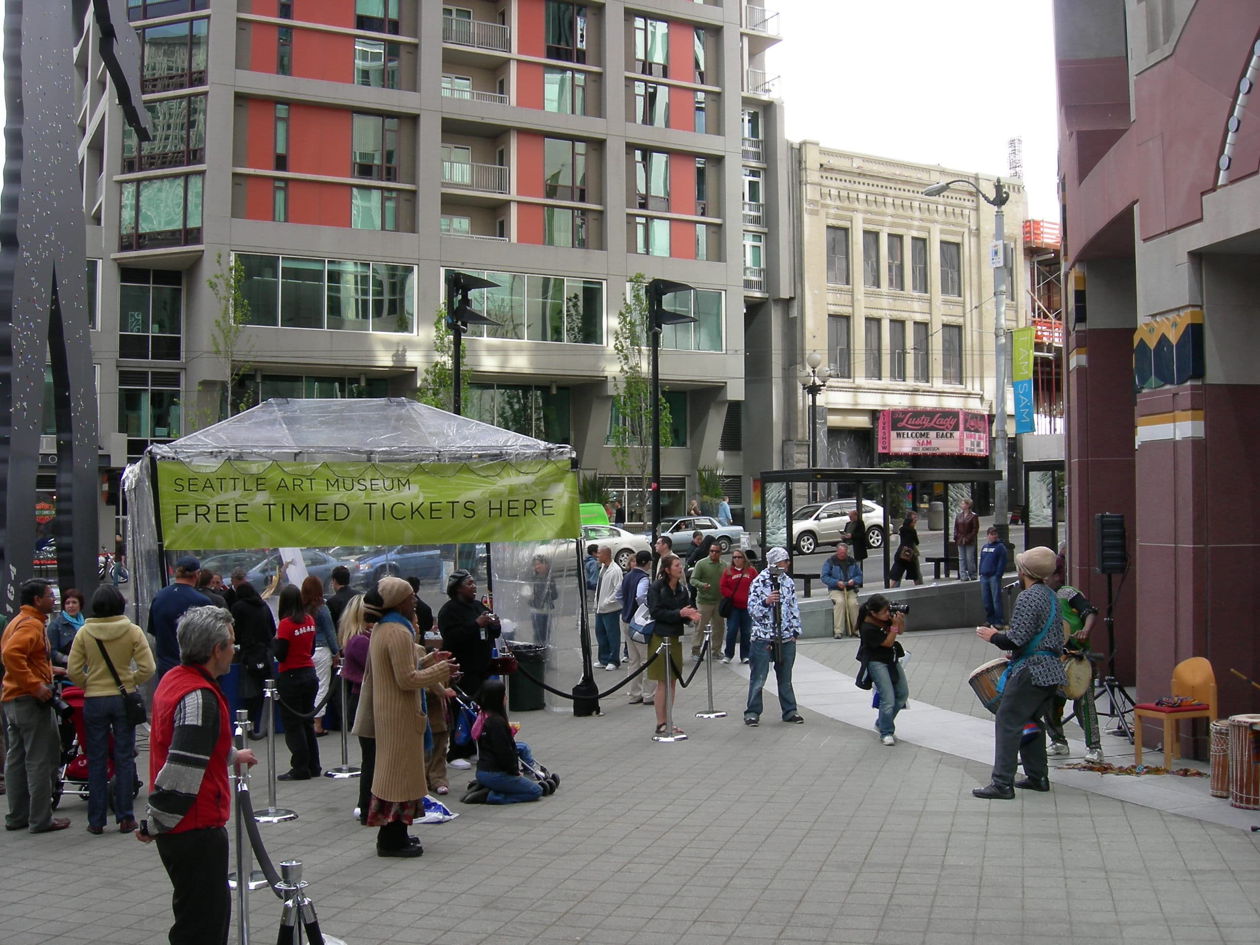 Real photograph of the Seven Seas Building in 2007: the tan-brick facade with the live pink Lusty Lady marquee reading Welcome Back SAM, across 1st Avenue from the Seattle Art Museum